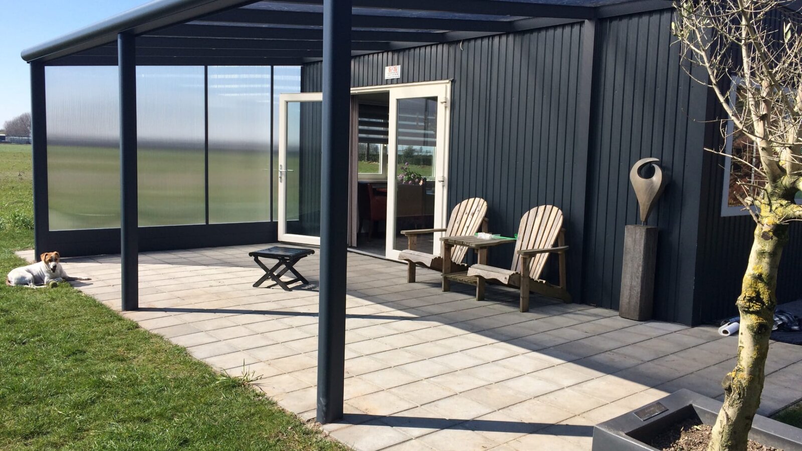 Glazen overkapping met donker aluminium frame en glazen zijwanden in een groene tuin